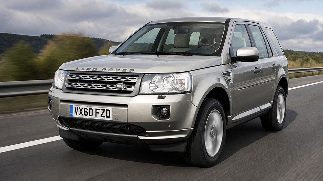 Carro SUV Land Rover cinza a circular numa estrada com vegetação e céu nublado ao fundo.