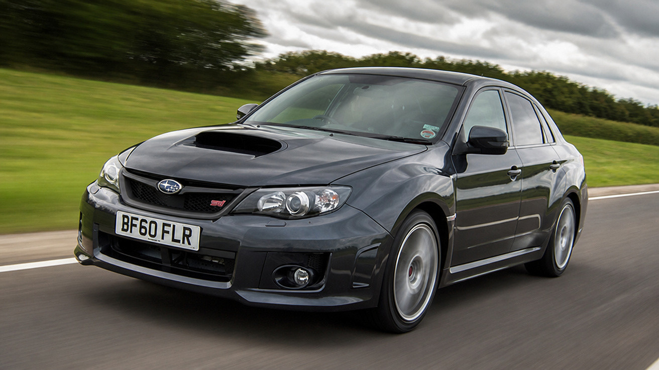 Carro desportivo Subaru WRX STI preto em movimento numa estrada com paisagem verde ao fundo.