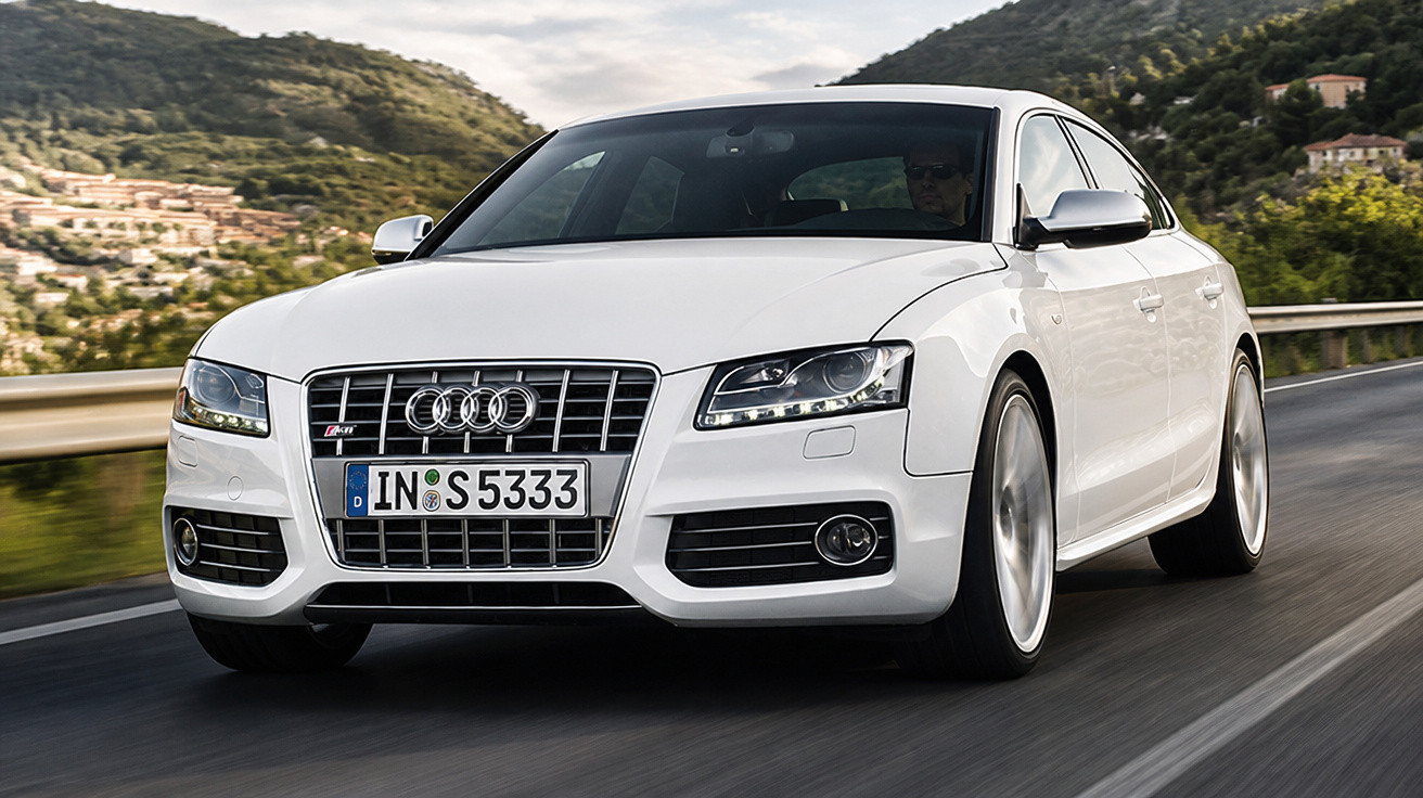 Carro Audi branco em movimento numa estrada rodeada de colinas verdes e casas ao fundo.