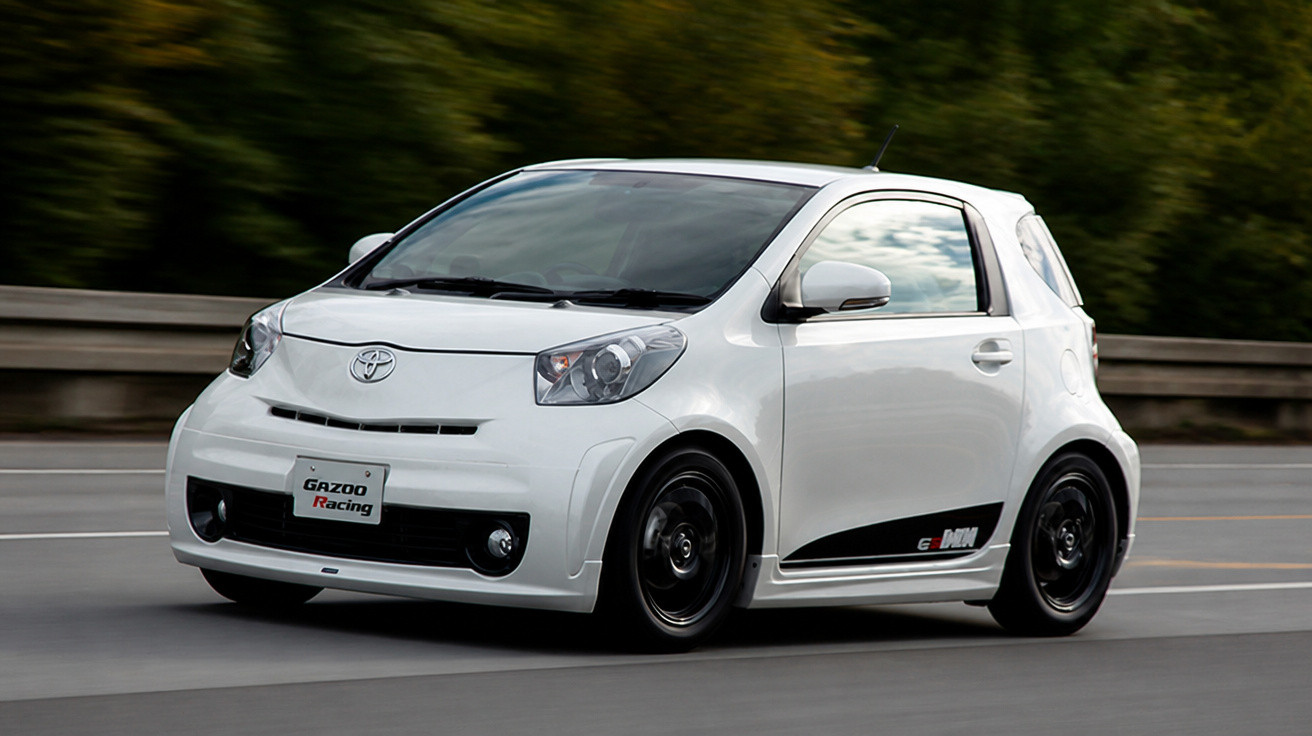 Carro compacto branco Toyota em movimento numa estrada com árvores ao fundo.