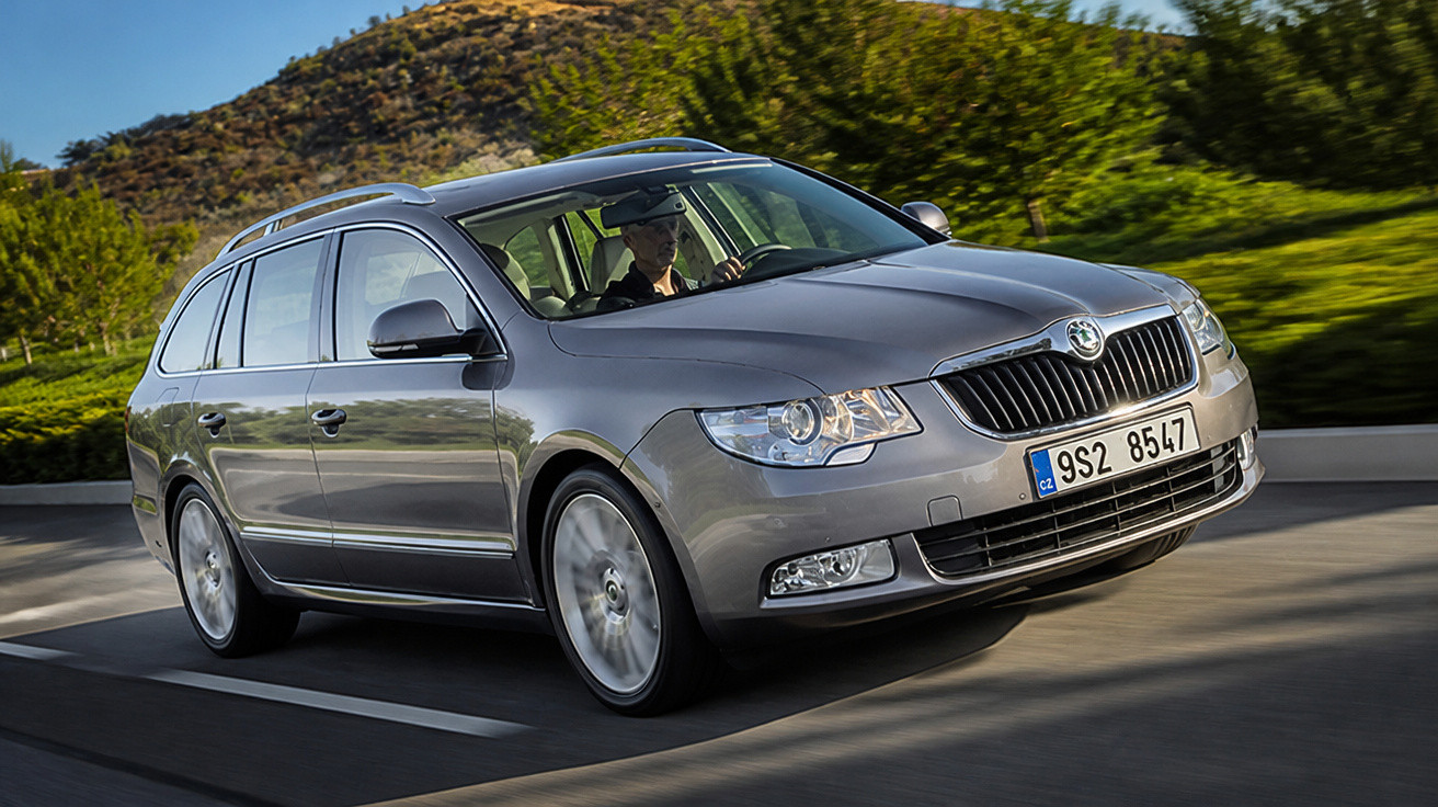 Carro familiar Skoda cinza em movimento numa estrada, com paisagem verde e céu azul ao fundo.