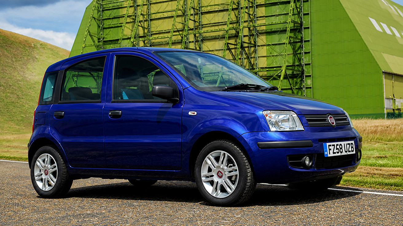 Carro Fiat Panda azul estacionado numa estrada com estrutura verde e colina ao fundo.