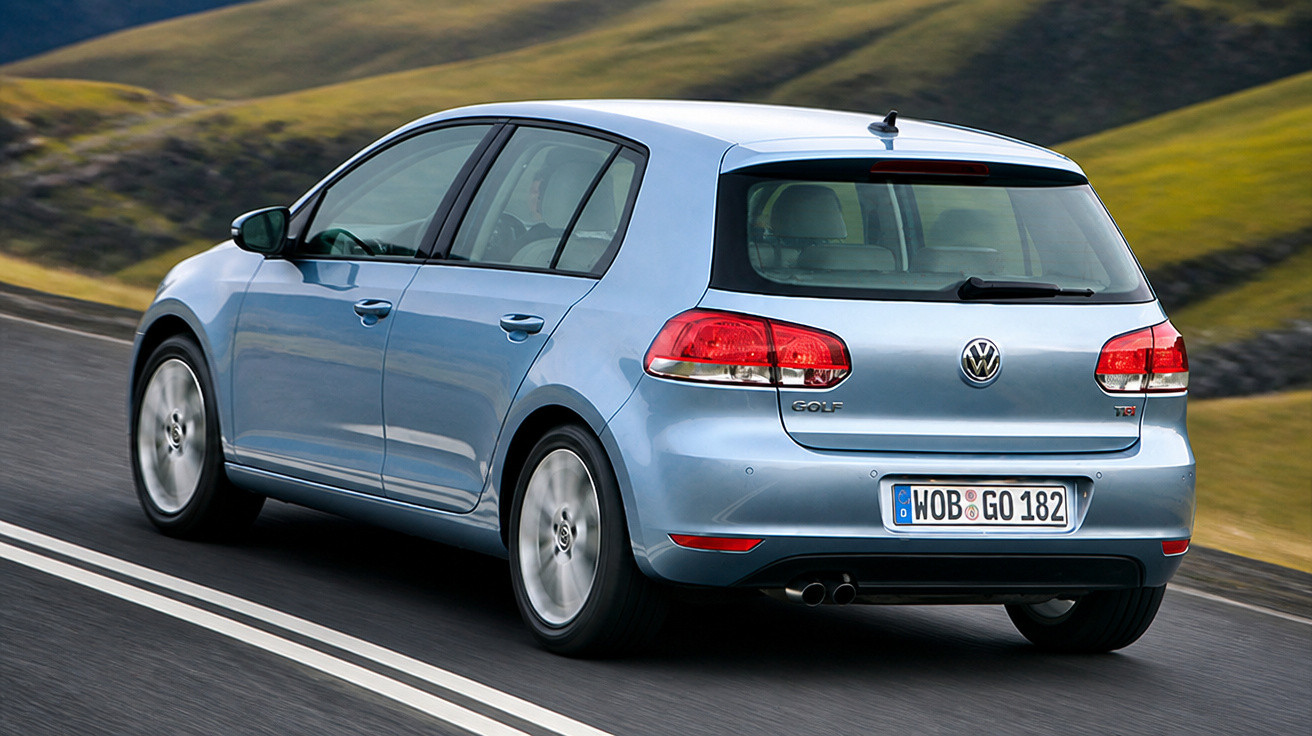 Automóvel Volkswagen Golf azul a circular numa estrada com relva e colinas ao fundo.