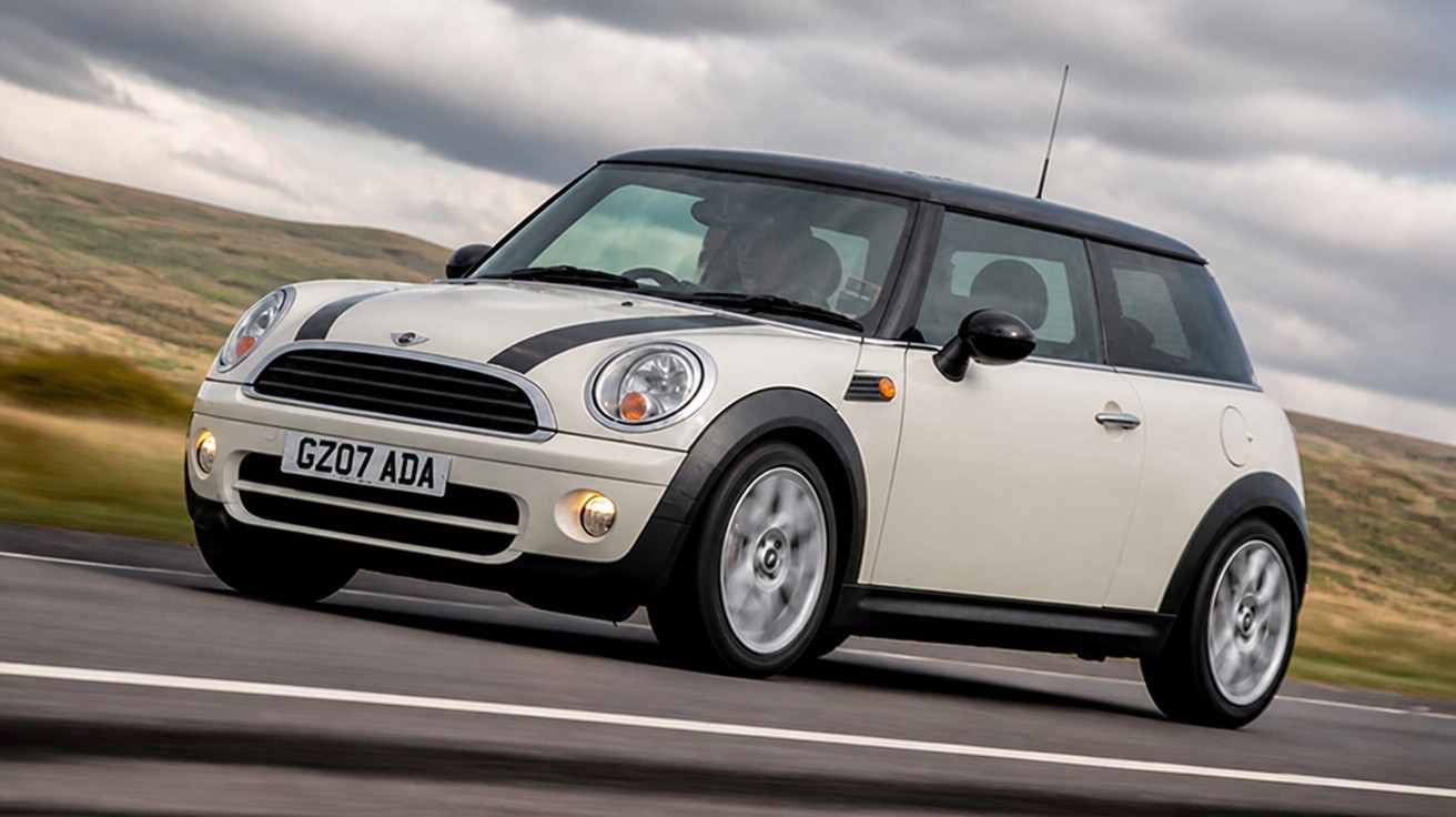 Carro Mini Cooper branco com teto preto a circular numa estrada sob céu nublado.