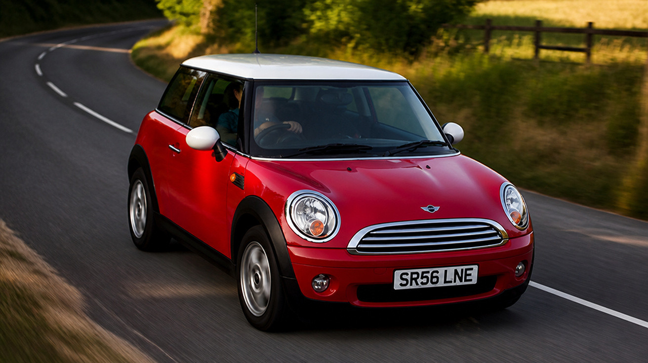 Carro Mini Cooper vermelho com tejadilho branco a conduzir numa estrada sinuosa rodeada de natureza.
