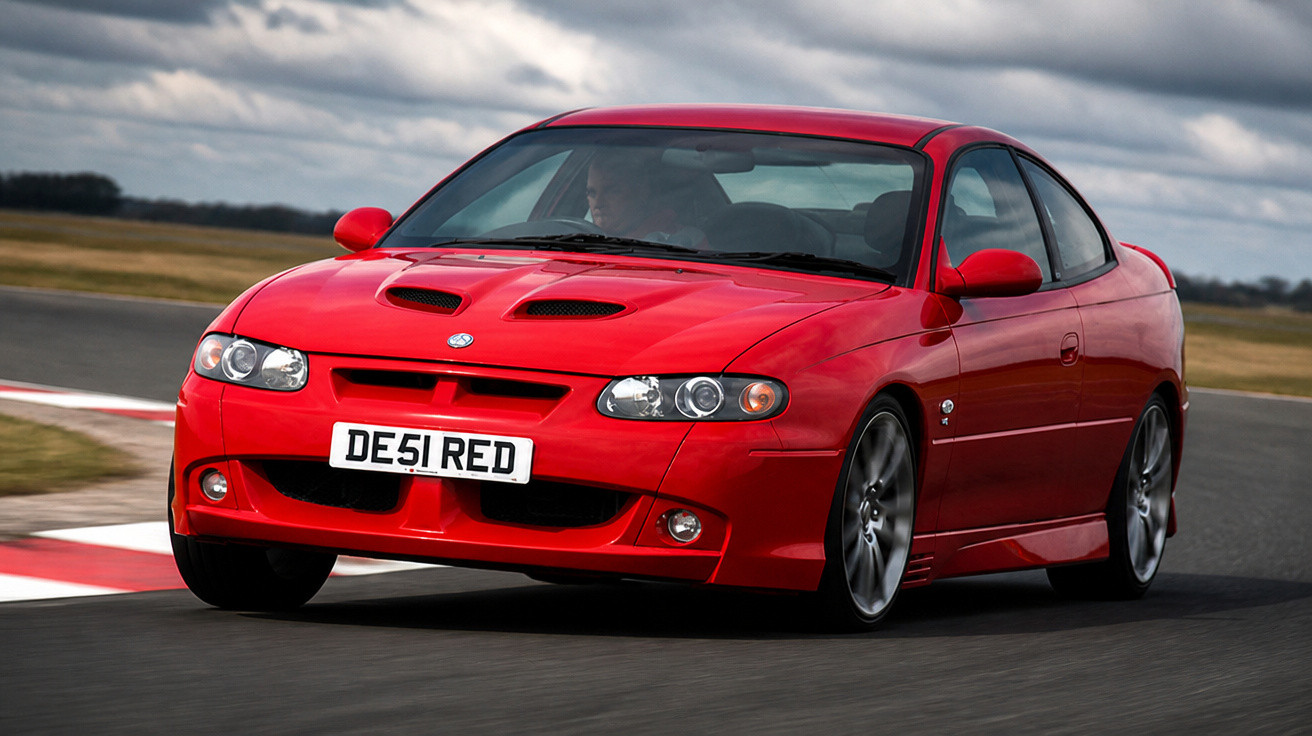 Carro desportivo vermelho a circular numa pista de corridas sob céu nublado.